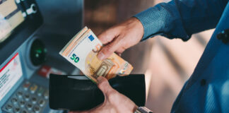Ein Mann hält ein Portemonnaie mit Euroscheinen in Händen und sortiert seine Scheine ein. Er trägt ein blaues Hemd und steht an einem Geldautomaten.