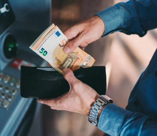 Ein Mann hält ein Portemonnaie mit Euroscheinen in Händen und sortiert seine Scheine ein. Er trägt ein blaues Hemd und steht an einem Geldautomaten.