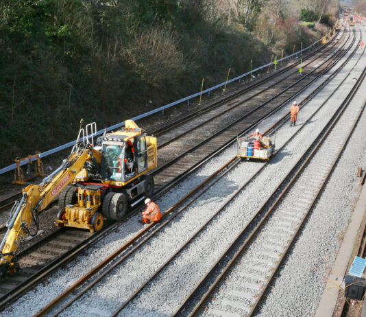 Bauarbeiter führen Arbeiten an einer Bahnstrecke durch. Die Strecke ist leer, kein Zug fährt mehr. Die Schienen werden repariert. Ein Kran und mehrere Arbeiter arbeiten auf der Baustelle an den Gleisen.