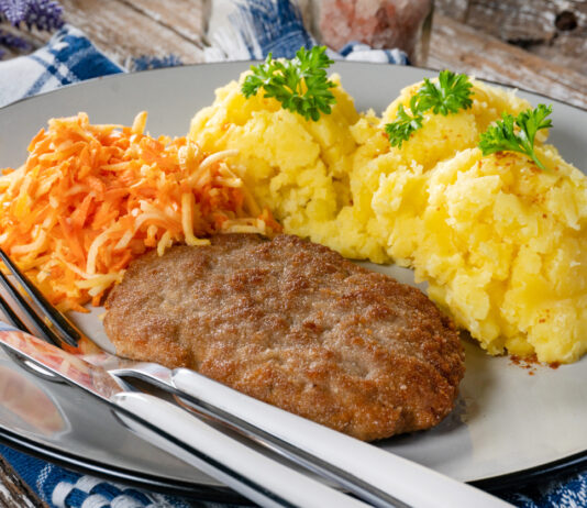 Eine Mahlzeit bestehend aus Kartoffelbrei, Karotten und Hackschnitzel liegt auf einem Teller mit Messer und Gabel. Der Kartoffelbrei ist garniert mit Petersilie.