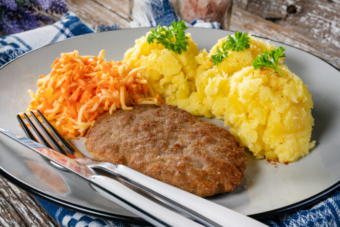 Eine Mahlzeit bestehend aus Kartoffelbrei, Karotten und Hackschnitzel liegt auf einem Teller mit Messer und Gabel