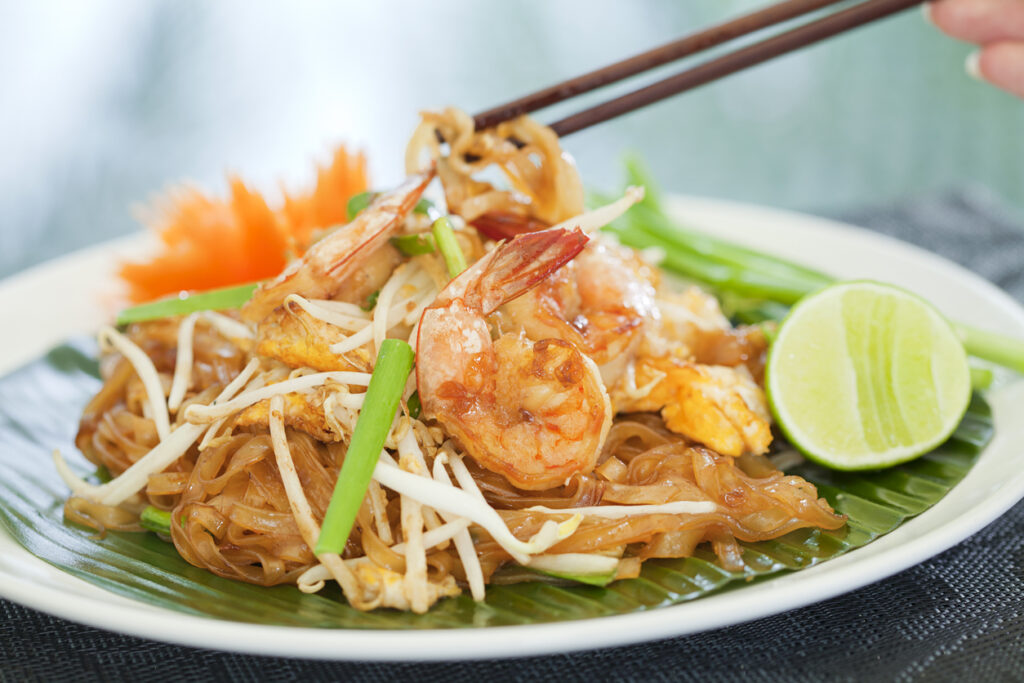 Pad Thai ist ein beliebtes thailändisches Gericht, das aus gebratenen Reisnudeln, Eiern, Tofu oder Garnelen und einer würzigen Tamarindensauce zubereitet wird. In Restaurants wird es oft mit frischen Limetten, Erdnüssen und Koriander serviert, um den Geschmack zu vervollkommnen.