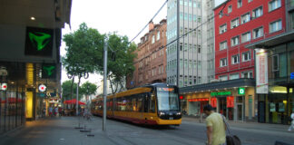 Die S-Bahn fährt durch die Innenstadt und hält an einer Haltestelle, an der Passanten warten.