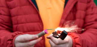 Ein Mann in einer roten Jacke hält einen kleinen Böller und zündet ihn mit einem Feuerzeug an.
