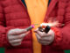 Ein Mann in einer roten Jacke hält einen kleinen Böller und zündet ihn mit einem Feuerzeug an.