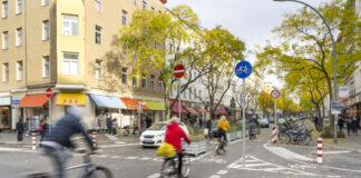 Fahrradfahrer und Autofahrer passieren eine Straße, an der Verkehrsschilder und Straßenschilder stehen