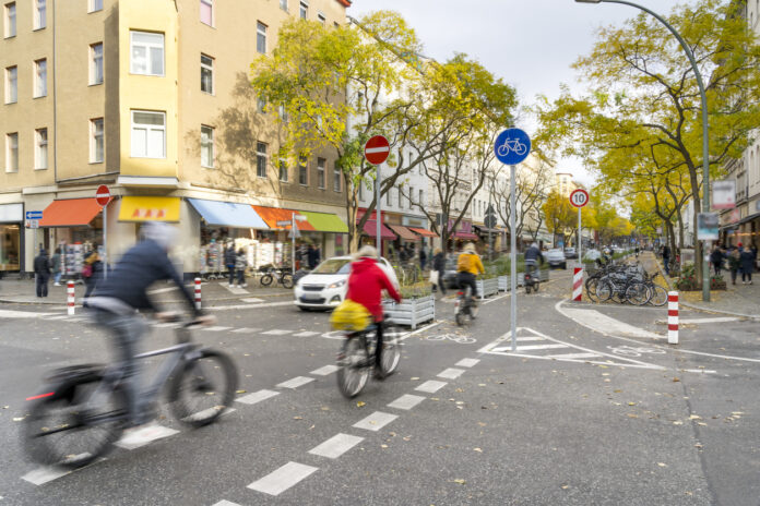 Fahrradfahrer und Autofahrer passieren eine Straße, an der Verkehrsschilder und Straßenschilder stehen