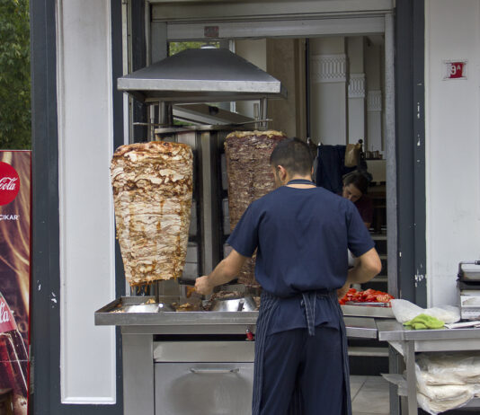 Ein Mitarbeiter steht an einem Dönerspieß und schneidet Fleisch für einen Döner ab. Im Hintergrund kann man in den Dönerladen hineinschauen.