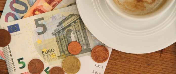 Auf einem Tisch neben einer Kaffeetasse liegen Münzen und ein 5 Euro Schein.