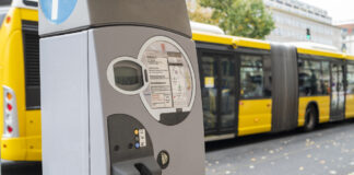 Ein grauer Parkautomat mit Münzeinwurfloch und Informationstafel steht an einer befahrenen Straße, die gerade ein gelb-schwarz lackierten Linienbus passiert, den man im Hintergrund sieht.