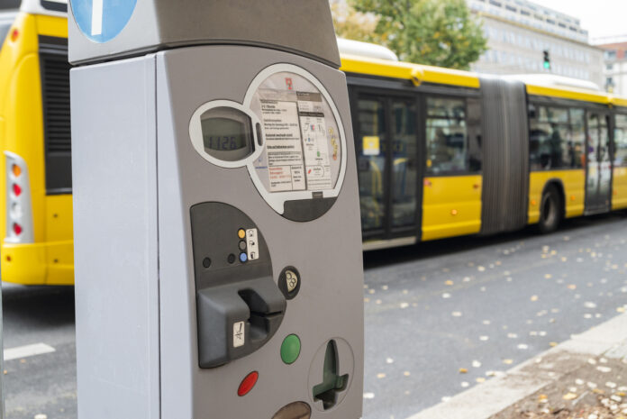Ein grauer Parkautomat mit Münzeinwurfloch und Informationstafel steht an einer befahrenen Straße, die gerade ein gelb-schwarz lackierten Linienbus passiert, den man im Hintergrund sieht.