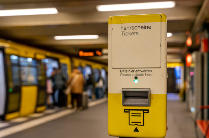 Im Vordergrund steht ein Automat, an dem man sein Ticket zur Kontrolle reinstecken muss. Im Hintergrund steigen Menschen in eine U-Bahn ein.