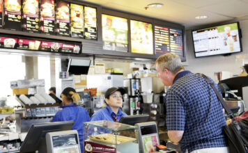 Eine Kasse in einem McDonald's. Das Personal nimmt eine Bestellung von einem Mann an. Im Hintergrund arbeiten die Mitarbeiter an der Zubereitung der Getränke und Speisen.