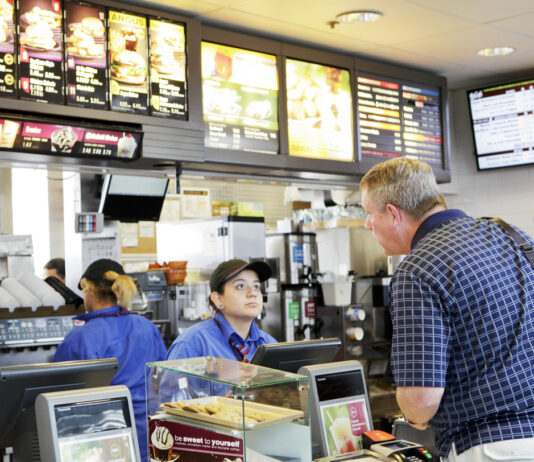 Eine Kasse in einem McDonald's. Das Personal nimmt eine Bestellung von einem Mann an. Im Hintergrund arbeiten die Mitarbeiter an der Zubereitung der Getränke und Speisen.