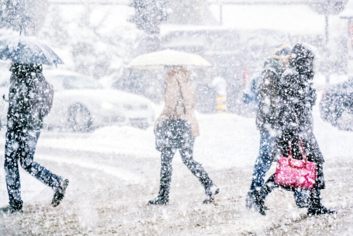 Es ist tiefster Winter in einer Großstadt. Einige Menschen laufen durch das Schneegestöber. Sie haben ihre Schirme aufgespannt. Im Hintergrund erkennt man einige Autos. Auch die Straßen sind zugeschneit.