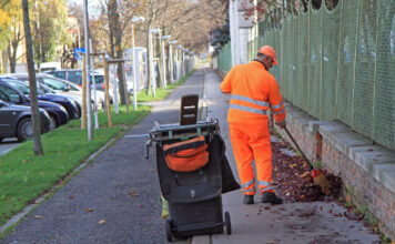 Ein Mann in oranger Arbeitskleidung fegt die Straße. Neben ihm steht eine Mülltonne.