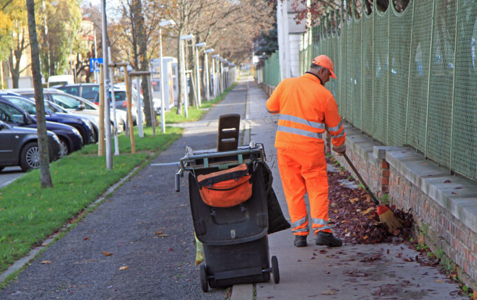 Ein Mann in oranger Arbeitskleidung fegt die Straße. Neben ihm steht eine Mülltonne.