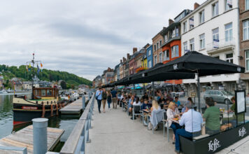 Touristen schlendern an einem Flussufer entlang. Andere sitzen in einem Cafe und essen und trinken.