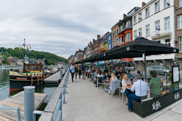 Touristen schlendern an einem Flussufer entlang. Andere sitzen in einem Cafe und essen und trinken.