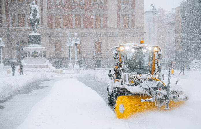 Ein Schneepflug räumt eine deutsche Straße, die hoch mit Schnee bedeckt ist. Man erkennt dies noch an den Stellen, an denen die Maschine noch nicht war. Im Hintergrund steht ein mit Schnee bedecktes Denkmal.