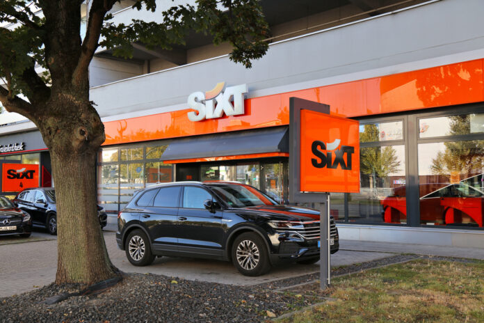 Ein Standort der Autovermietungsfirma Sixt und davor ein Auto neben einem Baum.