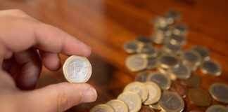 Eine Hand hält eine 1-Euro-Münze. Auf dem Tisch liegen viele 1-Euro-Münzen