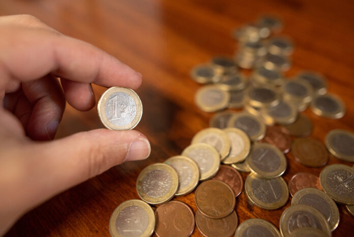 Eine Hand hält eine 1-Euro-Münze. Auf dem Tisch liegen viele 1-Euro-Münzen