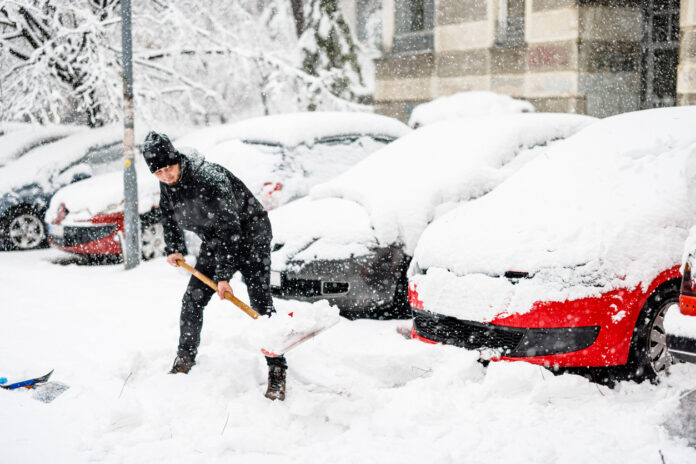 Es ist Winter in einer Großstadt. Die Gehwege und geparkten Autos sind komplett eingeschneit und kaum zu erkennen. Ein Mann mit einer dicken Winterjacke und Mütze schippt Schnee und versucht, den Weg freizumachen.