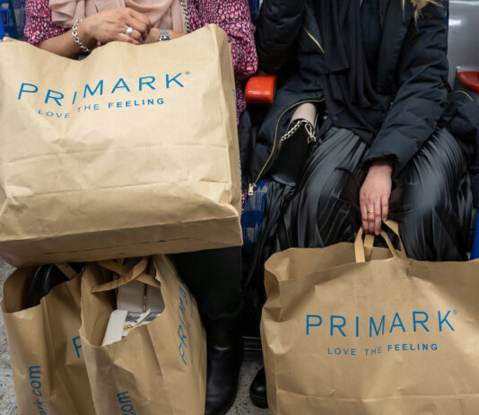 Frauen sitzen in der Straßenbahn und halten ihre Einkäufe vor sich. Diese befinden sich in Primark-Tüten aus Papier. Die Frauen sind elegant gekleidet.
