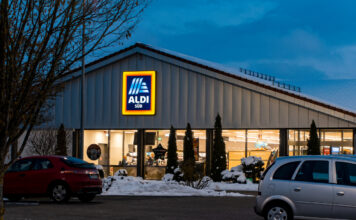 Eine Filiale von Aldi Süd steht in der Abenddämmerung. Es ist schon dunkel, daher ist bald Ladenschluss. Im Vordergrund auf dem Parkplatz stehen zwei Autos, und es liegt ein bisschen Schnee.