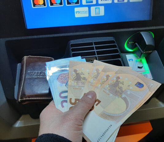 Ein Mann hebt Geld am Geldautomaten ab. In der Hand hält er verschiedene 50- und 20-Euro-Scheine, die er zu einem Fächer aufgereiht hat.