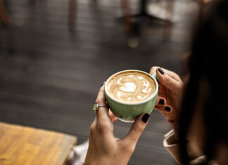 Eine unkenntliche Frau hält eine Tasse Kaffee mit Milch in der Hand. Die Tassse ist grün, der Kaffee frisch. sie sitzt offenbar in einem Café mit Holztischen und Holzboden.