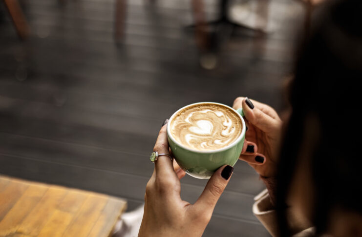 Eine unkenntliche Frau hält eine Tasse Kaffee mit Milch in der Hand. Die Tassse ist grün, der Kaffee frisch. sie sitzt offenbar in einem Café mit Holztischen und Holzboden.