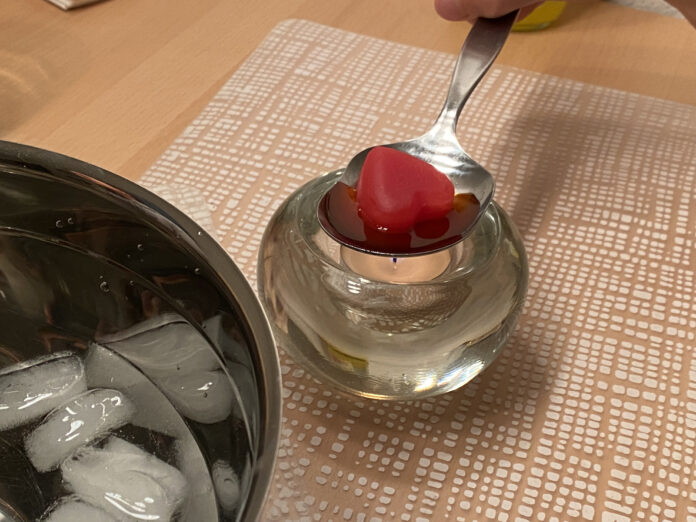 Ein rotes Wachsherz in einem Löffel neben einem Kübel mit Eiswürfeln.