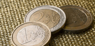 Zwei Ein-Euro-Stücke und ein Zwei-Euro-Stück liegen auf einer goldenen und strukturierten Oberfläche. Das Bargeld ist gold und silber und aufeinander gestapelt.