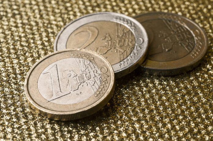 Zwei Ein-Euro-Stücke und ein Zwei-Euro-Stück liegen auf einer goldenen und strukturierten Oberfläche. Das Bargeld ist Gold und Silber und aufeinander gestapelt.