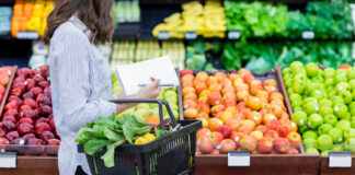 Eine Frau im Supermarkt trägt einen Korb, der voll ist mit Obst und Gemüse. Sie geht an den Auslagen vorbei, in denen sich verschiedene Sorten befinden.