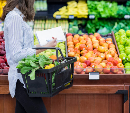 Eine Frau im Supermarkt trägt einen Korb, der voll ist mit Obst und Gemüse. Sie geht an den Auslagen vorbei, in denen sich verschiedene Sorten befinden.