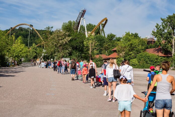 Eine lange Schlange von Menschen in einem Freizeitpark. Im Hintergrund erkennt man mehrere Achterbahnen, eingebettet zwischen grünen Bäumen und Büschen. Es scheint Sommer zu sein und viele Gäste des Freizeitparks tragen kurze Hosen.