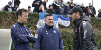 Die Verantwortlichen und Trainer sowie die Spieler des Karlsruher SC stehen zusammen in einer Beratung vor einem Fußballspiel. Sie haben die typische blau-weiße Mannschafts-Bekleidung an.