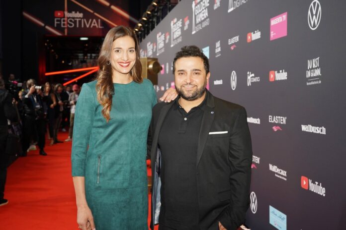 Sally steht neben ihrem Mann Murat Özcan bei einem öffentlichen Event auf dem roten Teppich vor einer Werbetafel