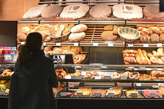 Eine Kundin steht in einer Bäckerei vor einer Auslage von verschiedenen Backwaren wie Brot, Brötchen und Süßgebäck und wartet auf die Bedienung, während sie sich das Brot anschaut.