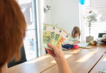 Eine Frau hält Geldscheine in ihrer Hand, im Esszimmer hoch, und zeigt sie ihrem Kind, welches am Tisch sitzt und Hausaufgaben macht.