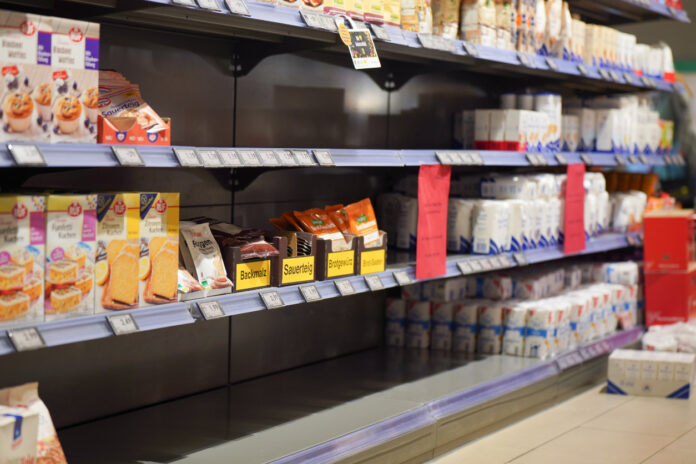 Regale in einem Supermarkt. Viele Produkte sind ausverkauft, weshalb zum Teil leere Regale zu sehen sind. Es stehen noch Kuchenbackmischungen und Mehl sowie einige weitere Artikel in den Regalen.