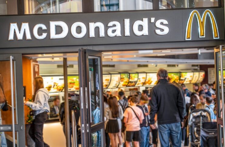 Eine überfüllte McDonald's Filiale. Sowohl vor allen Schaltern als auch vor der Filiale selbst binden sich lange Schlangen. Wegen einer Maßnahme gehen die McDonald's-Kunden auf die Barrikaden.