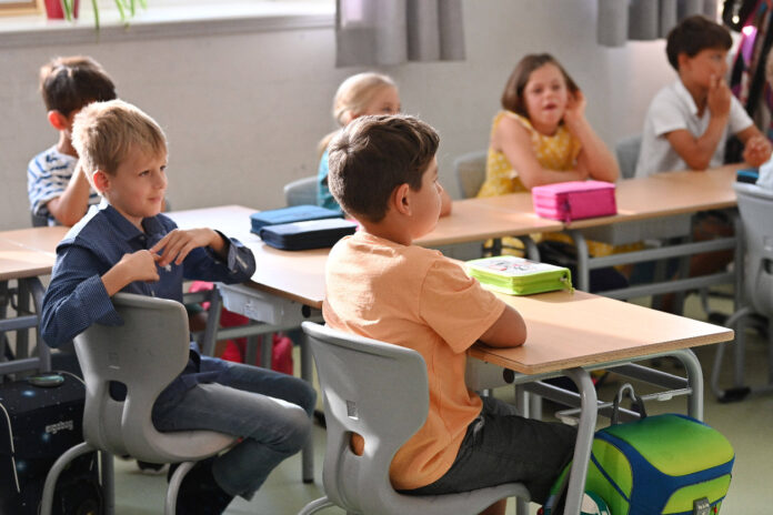 Mehrere Kinder sitzen in ihrer Klasse an ihren Tischen in der Schule und hören ihrem Lehrer oder ihrer Lehrerin mehr oder weniger aufmerksam zu.