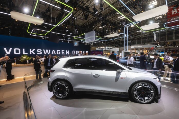 Ein weißes Elektroauto von Volkswagen wird bei einer großen Automesse präsentiert. Das Auto ist glänzend, neu, hochwertig und lockt zahlreiche Besucher an.