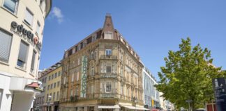 Mehrere Personen sind in der Karlsruher Innenstadt unterwegs. Die Passanten überqueren die Straße an einem großen Gebäude an der Ecke. Die historische Architektur ist im Sonnenlicht der Stadt gut zu erkennen.