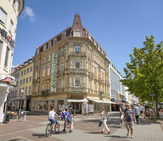 Mehrere Personen sind in der Karlsruher Innenstadt unterwegs. Die Passanten überqueren die Straße an einem großen Gebäude an der Ecke. Die historische Architektur ist im Sonnenlicht der Stadt gut zu erkennen.
