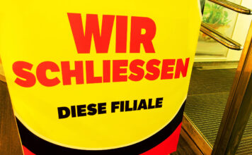 In einer Filiale in einer Modekette steht ein Schild mit der Aufschrift "Wir schließen diese Filiale". Das Schild verweist auf einen Schlussverkauf und günstige Preise. Die Produkte sind nur noch im begrenzten Maße verfügbar.
